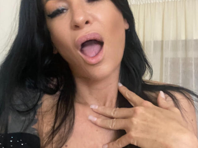 AndraD has black hair, open mouth, tongue slightly out, and her hand touching her neck. She has visible makeup and is wearing a black top with small studs.
