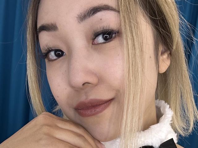 RenyLime's face, blonde hair, brown eyes, light skin, subtle makeup, dark lipstick, slight smile, right hand under chin, blue curtain in background, white furry collar on black jacket.