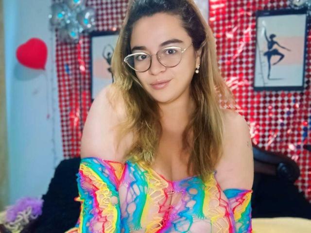 NaugthyCurvy, com óculos, cabelo loiro ondulado e uma blusa colorida, com um ombro de fora e babados, mostrando os seios, sorrindo para a câmera..
