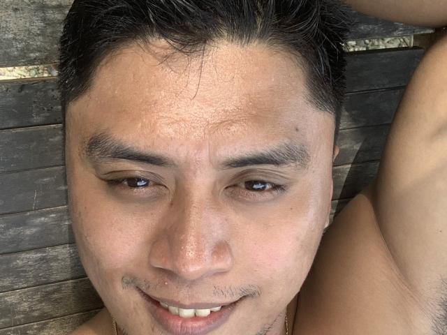 ImJiro's face, shirtless, with black hair, light brown skin, small mustache, and smile, arm raised, showing armpit hair, lying on wooden surface.