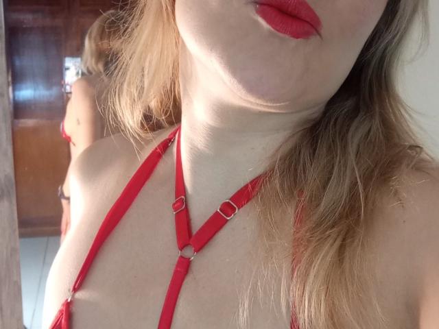 MadissonS, avec des lèvres rouges et de la lingerie fine rouge, montrant ses seins dans un reflet de miroir. Ses cheveux blonds sont lâchés, et elle a un teint clair..
