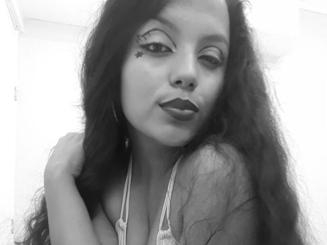 KonyBella with long, wavy dark hair, wearing a halter top, winking, has a star tattoo under her right eye, and dark lipstick, holding her hair with one hand.