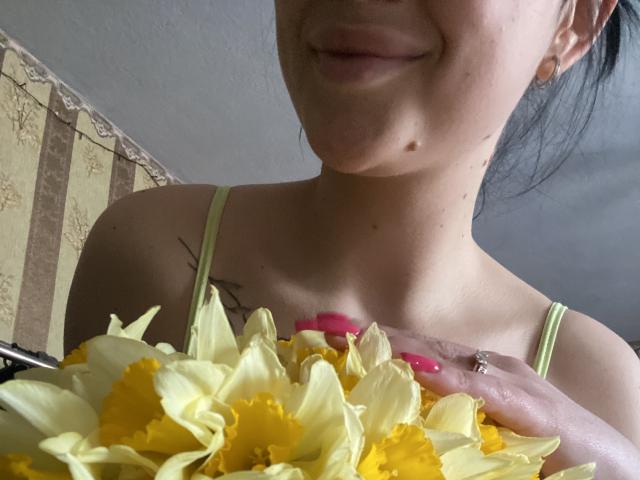 Olieri with yellow and white daffodils, holding them near her chest, has pink nails, a small mole on her chin, and a tattoo on her right collarbone.
