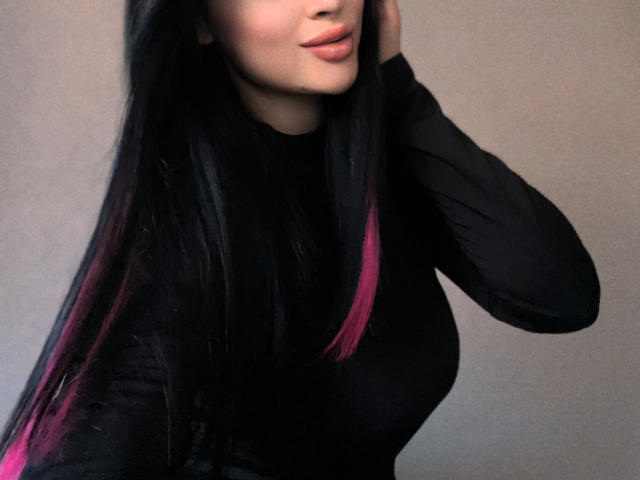 Olieri with long black hair, pink tips, wearing a black long-sleeve shirt, has her hand in her hair, showing her pouty lips and her curvy, ample tits.