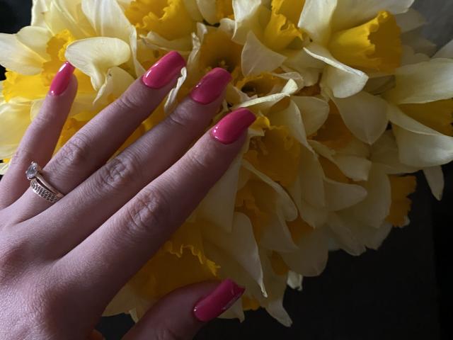 Olieri's hand with pink glossy nails, yellow daisy, and diamond ring, touching petals. Her fingers are slightly curled, showing off her shiny nails and sparkly ring.