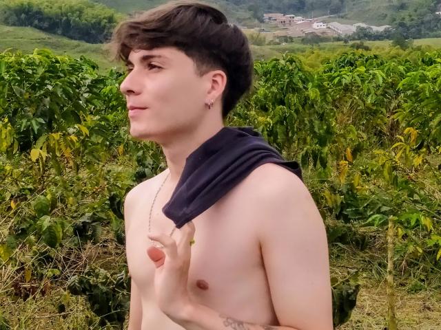 Jack Rouse, sin camisa y con el pelo corto y castaño, sostiene una camisa negra sobre su hombro. Tiene una complexión delgada, pezones pequeños y un tatuaje en el brazo derecho..