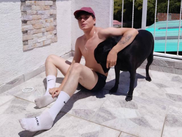 JackRouse, sin camisa, con pantalones cortos negros, calcetines blancos, gorra roja, sentado en el suelo de baldosas, un perro negro a su derecha, tatuaje en el brazo izquierdo, piscina al fondo, día soleado..