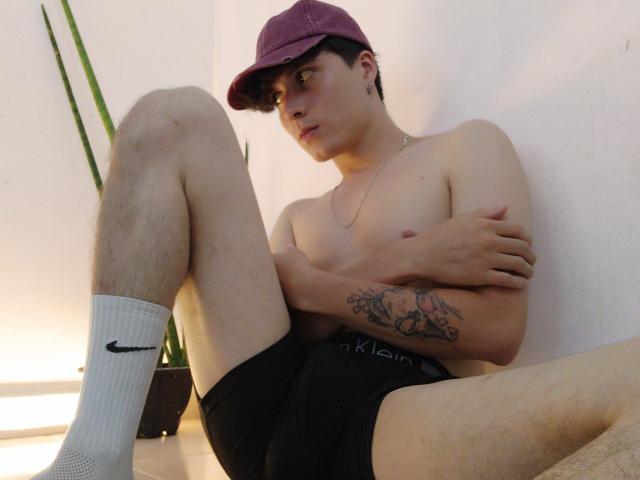 JackRouse, sin camisa, con pantalones cortos negros, calcetines blancos con el logo de Nike, gorra morada, tatuaje en el brazo, sentado con las piernas separadas, brazos cruzados, mirando hacia un lado, piel pálida, cabello negro corto..