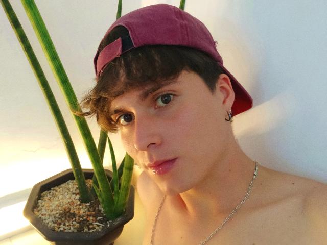 JackRouse, con cabello castaño corto, usando una gorra morada puesta al revés, pequeños aretes y un collar de plata, mirando a la cámara con una ligera sonrisa, al lado de una planta verde..