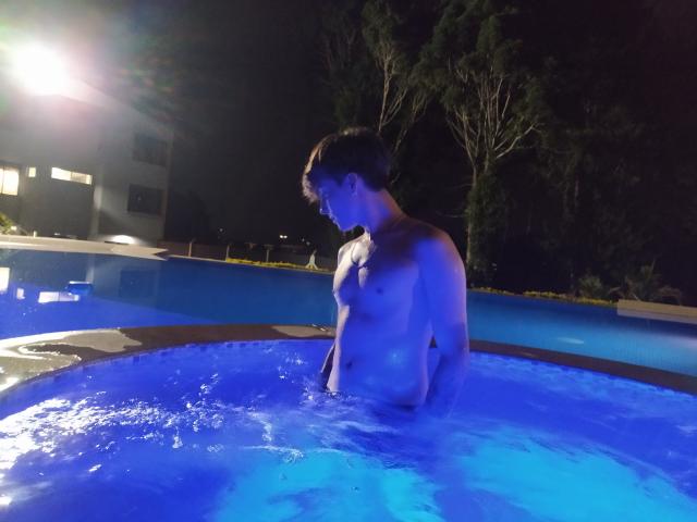 JackRouse, sin camisa, está de pie en una bañera de hidromasaje azul brillante, con el agua hasta las nalgas, mirando hacia abajo, con el pelo corto y castaño, de noche, con poca luz, y árboles en el fondo..