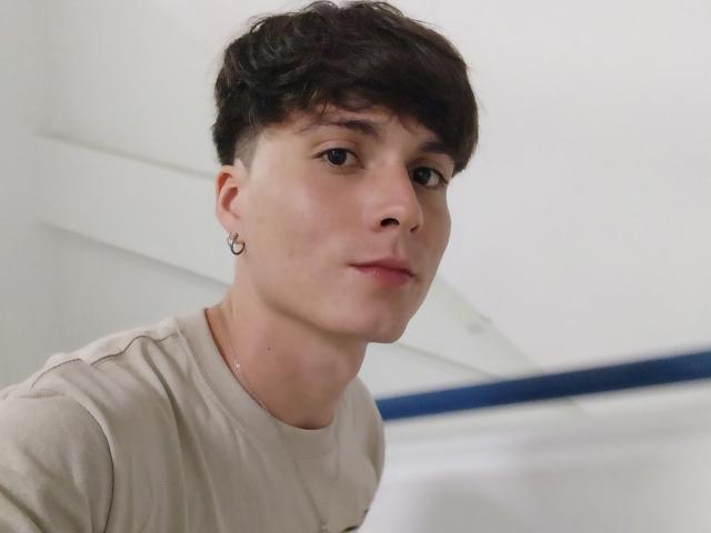 JackRouse, un joven con cabello oscuro y corto, un pequeño aro en la oreja, piel clara, una sonrisa sutil, vistiendo una camisa beige, con un fondo ligeramente borroso..