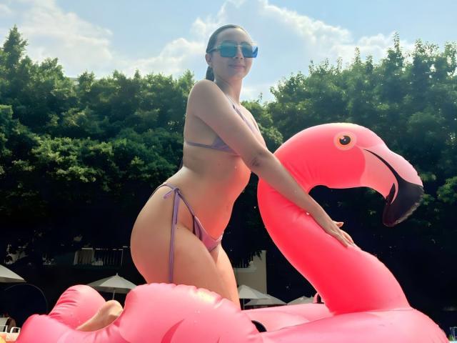 MajoGomez with pink flamingo inflatable, wearing purple bikini, her ass and tits noticeable, blue sunglasses, standing on inflatable, sunny day, green trees in background.