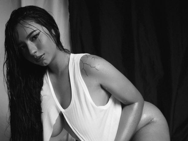 Mia Suarez, les cheveux mouillés, portant un débardeur blanc qui expose ses seins, et montrant un tatouage sur son épaule, penchée en avant, avec des gouttes sur sa peau brillante..