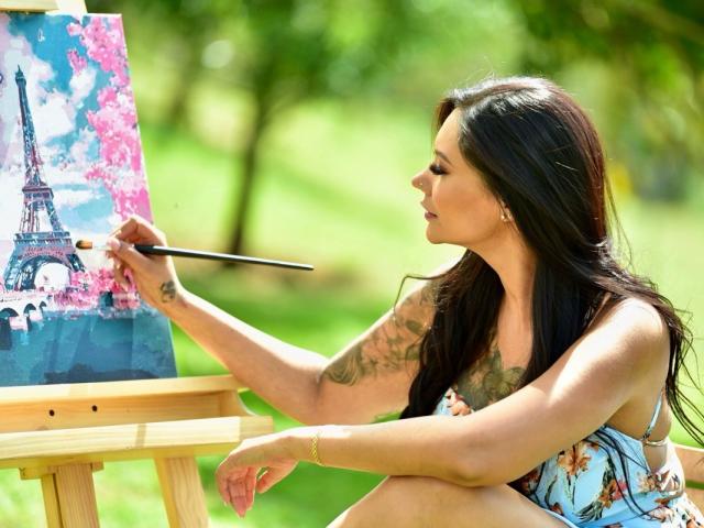 AlessandraMalory with long black hair, tattooed arm, floral dress, painting Eiffel Tower on canvas with brush, sitting outdoors, holding wooden easel, sunlight on green grass.