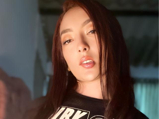 SaraCorny69 with light brown skin, dark brown hair, and slightly parted pink lips, wearing a black mesh top, looking seductively into the camera with a soft, glowing face.