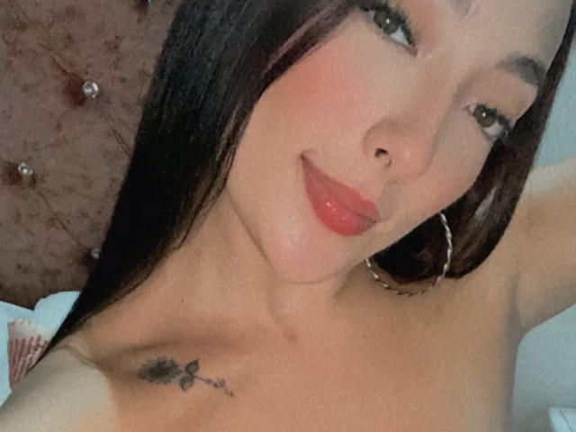 SaraCorny69 has long black hair, red lipstick, and a small tattoo under her right boob. She's wearing large hoop earrings and smiling softly at the camera.