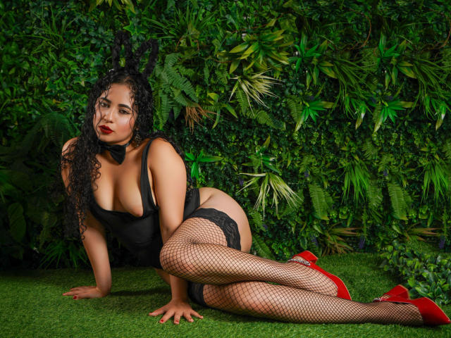 PerfectLyly, con cabello negro rizado, lápiz labial rojo, orejas de conejo negras, lencería negra, medias de red y tacones rojos, sobre césped verde, mostrando sus grandes pechos y glúteos..