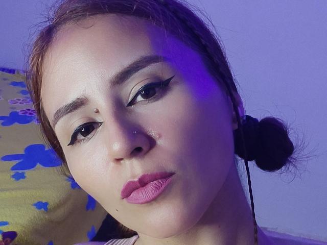 HannaHouse's face with brown hair in buns, pink lipstick, black eyeliner, nose piercing, and slight bruise on right cheek, under purple lighting, looking serious.