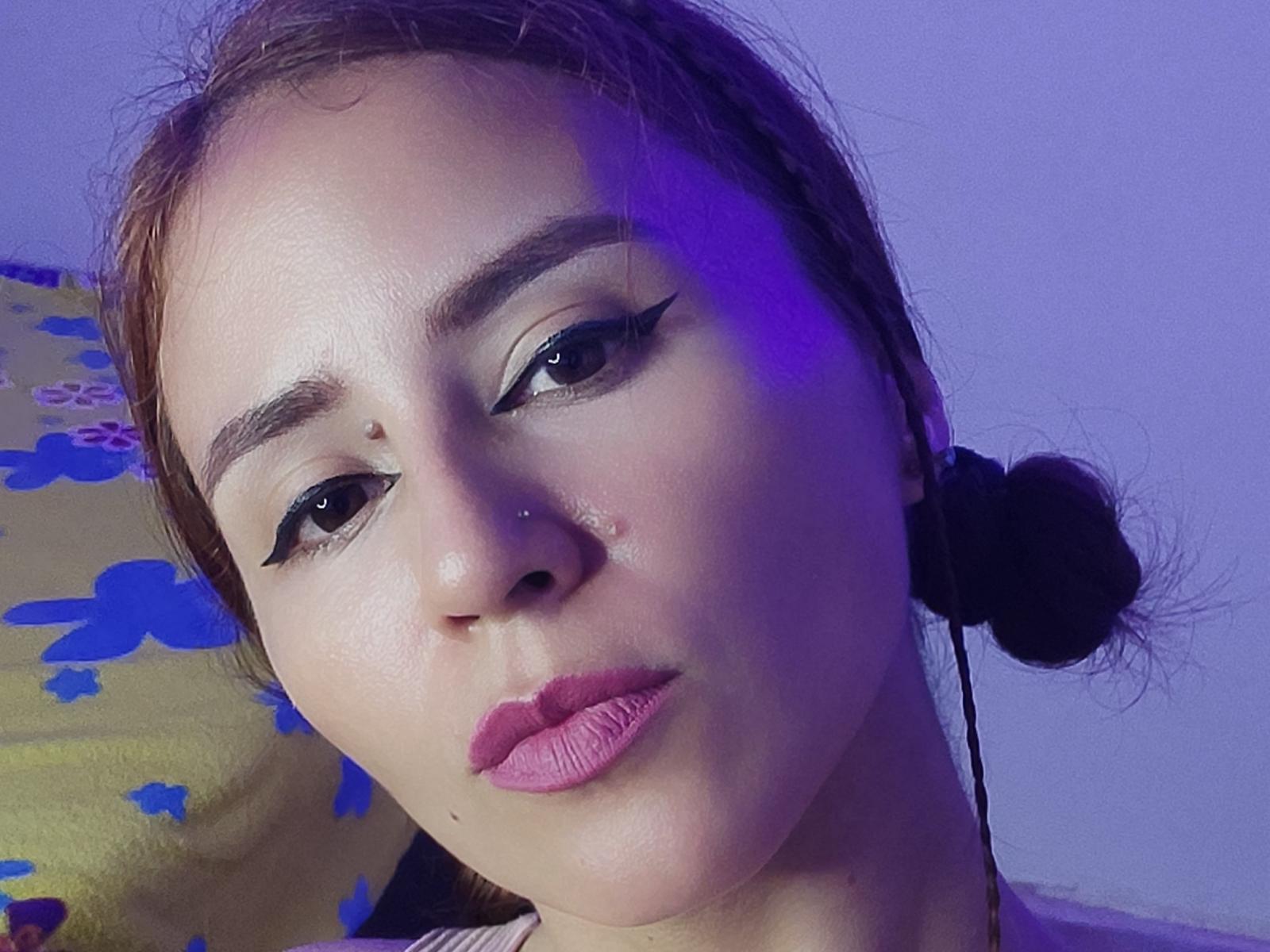 El rostro de HannaHouse, con cabello castaño recogido en moños, lápiz labial rosa, delineador de ojos negro, perforación en la nariz y un ligero moretón en la mejilla derecha, bajo una iluminación púrpura, con una expresión seria..