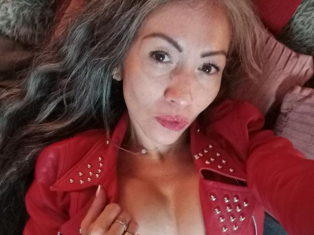 NicoletteShea with long gray hair, red studded jacket, and pink lipstick, looking at camera, hand on chest, showing cleavage, wearing necklace and ring, lying down.