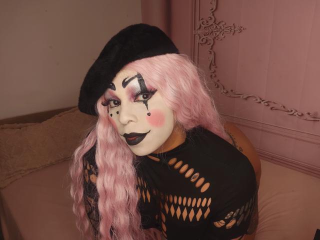 Chili with pink hair and black face paint is kneeling on a bed, wearing a black fishnet top and beret.