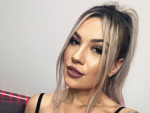 ZoeyMarie with blonde ponytail, dark eyebrows, full lips with mauve lipstick, wearing black strap top, subtle makeup, slight smile, light skin, small gold necklace, soft lighting.