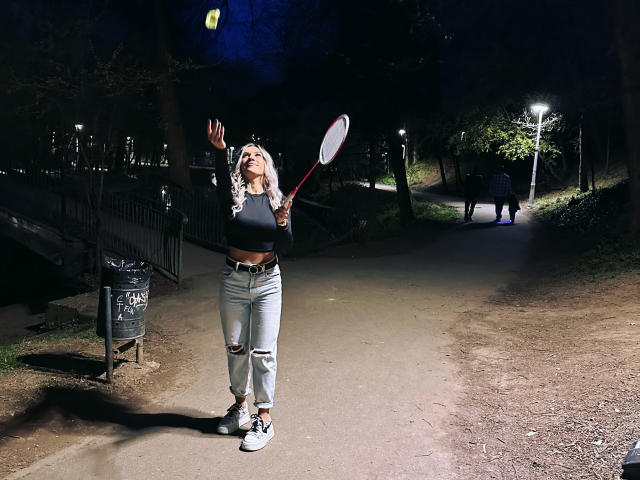 ZoeyMarie mit blonden Haaren, einem schwarzen Crop-Top, zerrissenen Jeans und weißen Sneakers, die einen Tennisracket hält, Nacht, Straße, Arm angehoben, leuchtender gelber Ball vor dunklem Hintergrund..
