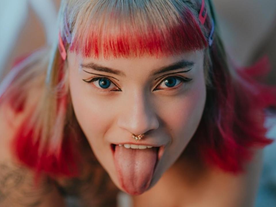 Darshelle with blonde and red-dyed hair, blue eyes, and a nose ring, sticking her tongue out, showing tattoos on her arm. She's looking directly at the camera.