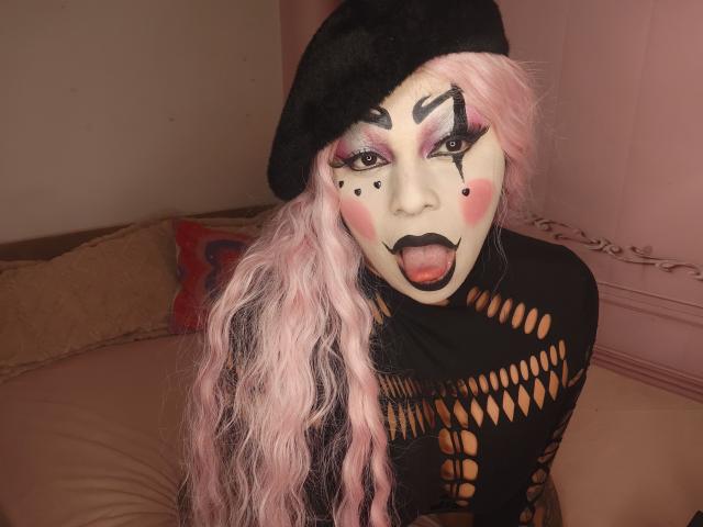 Chili with long pink hair, black beanie, and fishnet top, tongue out, makeup with black dots, leaning forward, sexy expression.