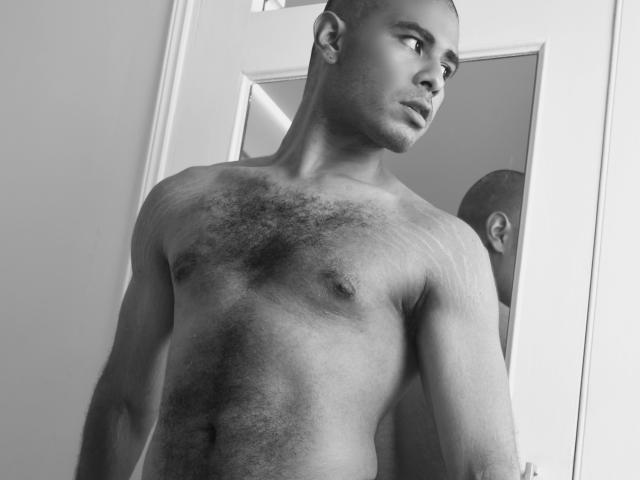 JhonOud, a muscular, shirtless black dude with a hairy chest, stands in a mirror's reflection, looking to the side. His flaccid, uncircumcised penis is not visible.