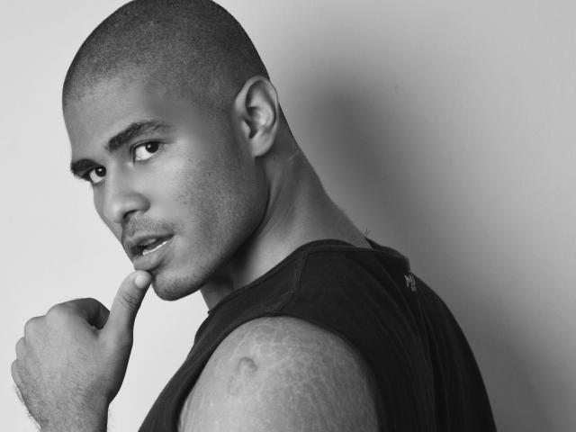 JhonOud has a shaved head, dark skin, and a muscular arm with tattoos. He's biting his finger, looking sideways with a serious expression in a black sleeveless shirt.