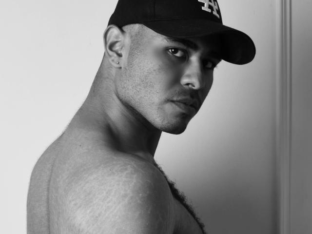 Black-and-white photo of JhonOud, shirtless with a baseball cap, looking over shoulder, showing a strong, hairy chest, and a piercing in his right ear. JhonOud's face is serious.
