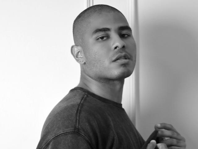 JhonOud with a shaved head, dark shirt, and earring, looking slightly to the side, holding something near his chest, in a black-and-white photo.