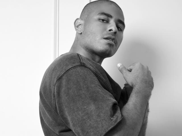 JhonOud, bald with a shaved head, wearing a dark, slightly textured shirt, looking over his shoulder, holding his shirt collar with a serious, slightly curious expression. Black-and-white photo.