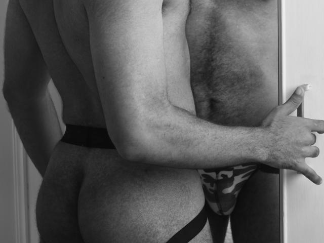 Black-and-white photo of JhonOud with a hairy chest, wearing a leopard print jockstrap, standing sideways, touching a mirror with his right hand, showing his bare ass.