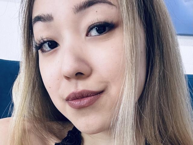 RenyLime's face with blonde hair, blue eyes, and dark eyeliner. She has full, pinkish-brown lips and a slight smile. Her skin is light with a few small moles.
