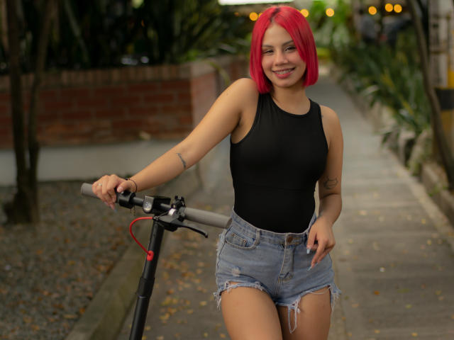 SussyGray, con i capelli rosa acceso, una canottiera nera senza maniche e pantaloncini di jeans sfilacciati, sorride mentre tiene una bicicletta su un marciapiede, mostrando un tatuaggio sul braccio destro..