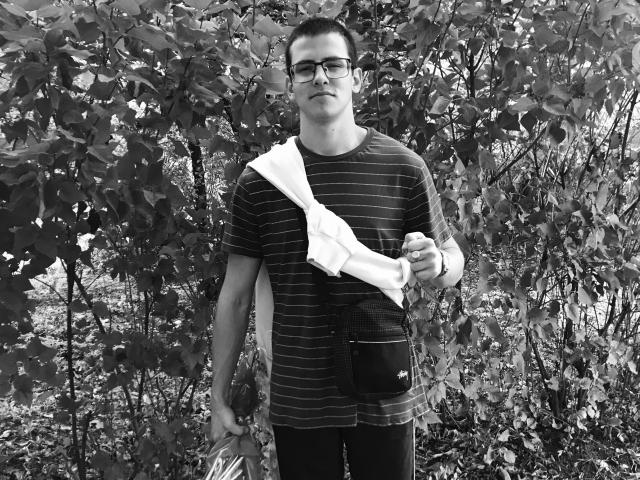 Black-and-white photo of StockingBoy with glasses, striped shirt, white bag, black pouch, holding a bottle, standing in leafy background, looking slightly annoyed. Short dark hair.
