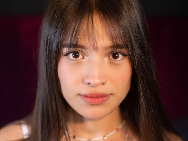 MelodySwet has brown eyes, straight brown hair with bangs, and pink lips. She's wearing a white tank top and a necklace. Her skin is fair. Her tits are small.