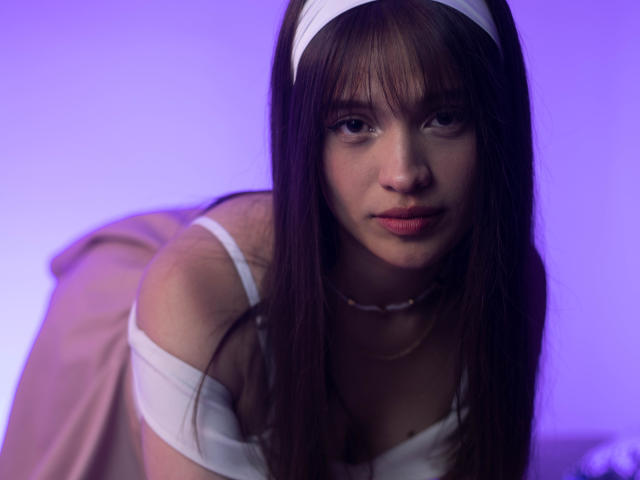 MelodySwet has long brown hair, a white headband, and is wearing a white top that's off-shoulder, looking down with a serious expression. She's in soft purple lighting.