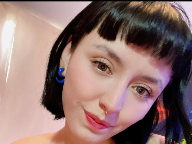 SaraleePoison hat kurze, schwarze Haare, blaue Ohrringe, eine Nasenring, ein leichtes Lächeln und rosafarbenen Lippenstift, braune Augen und ein süßes, süßes Gesicht..