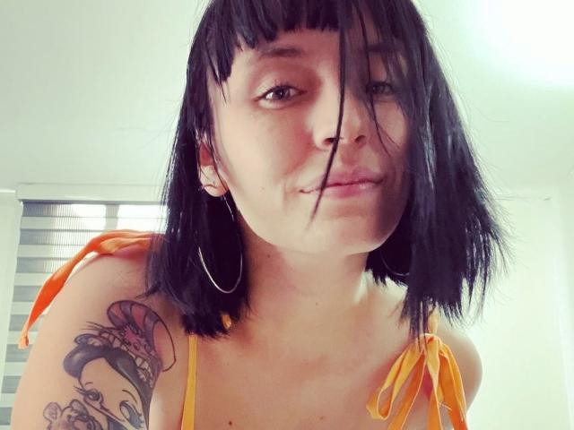 Saralee Poison, mit schwarzen Haaren, trägt ein gelbes Oberteil, große Ohrringe und hat ein Tattoo an ihrer rechten Schulter. Ihre Nase ist leicht hoch und eine Strähne ihres Haares bedeckt ihr Auge..