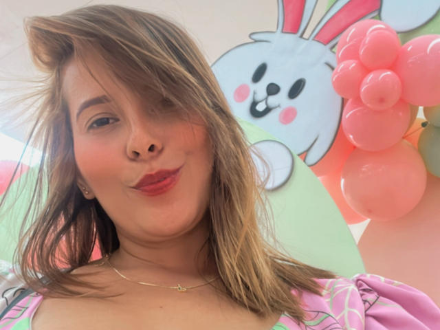 Sarita Velez, con i capelli castano chiaro, il rossetto rosa e un sorriso adorabile, indossa una maglietta rosa e bianca, ed è in posa davanti a decorazioni a forma di coniglio e a un palloncino rosa..