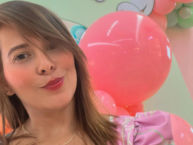 SaritaVelez with brown hair, pink lips, and a cute smile, holding pink balloons, wearing a pink and purple top, gold necklace, and looking slightly to the side.