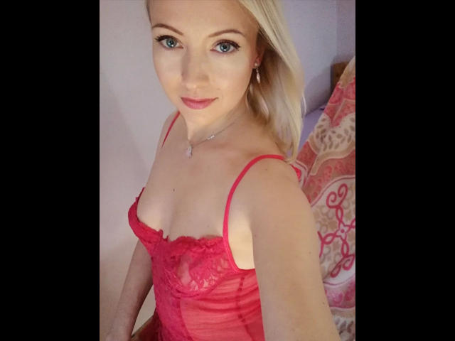 SusiPrivat224-hot with blonde hair, blue eyes, and red lace lingerie, showing her small tits and cute smile in a selfie. She's wearing a red spaghetti strap top.