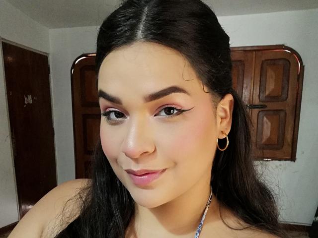 Antonela, avec de longs cheveux noirs, des yeux marron, un fard à paupières rose, du rouge à lèvres rose, des boucles d'oreilles cerceaux et un sourire discret, porte un haut à encolure plongeante, révélant un visage mignon et séduisant..