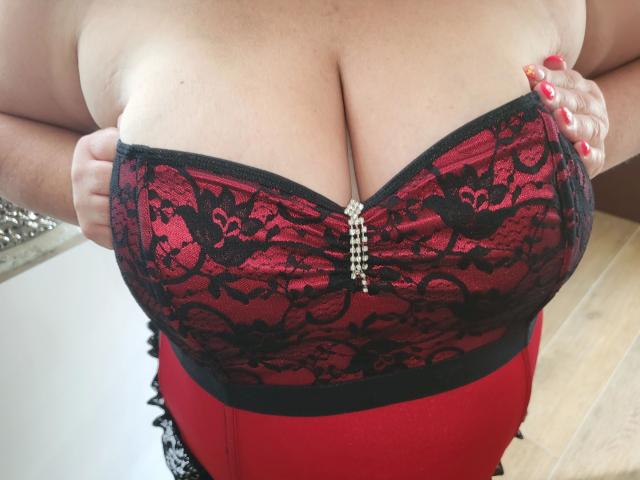 REALAMATEUR-hot's big boobs in a red and black lace bustier, hands pulling down the top to show cleavage, red nails, wood floor, and a small diamond button in center.