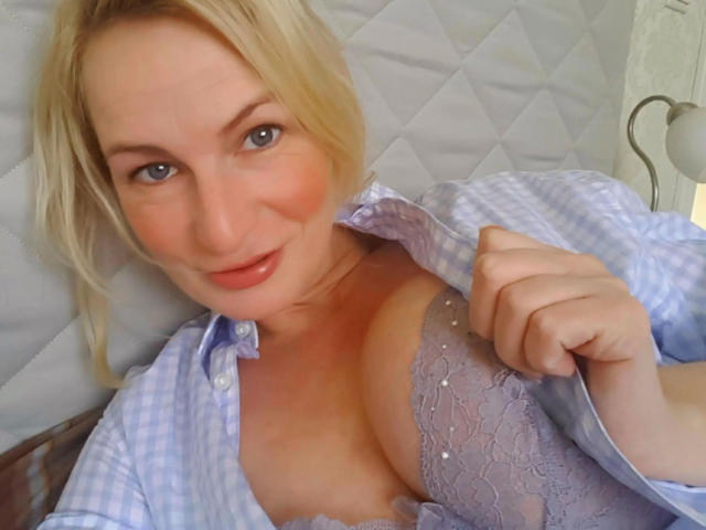 Blonde BAISE-MOI with blue eyes, slight smile, wearing an unbuttoned checkered shirt, exposing her lace-covered tits. She's lying down, holding her shirt open.