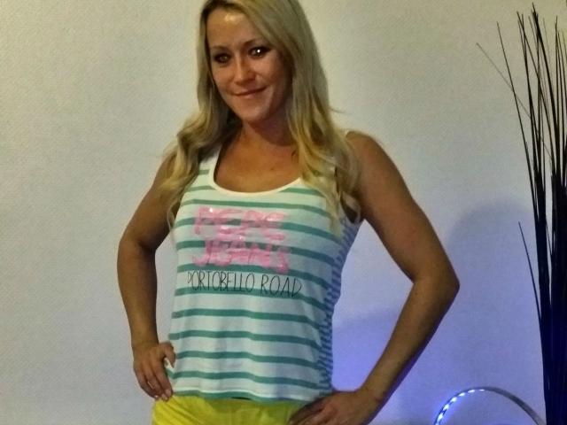 Blonde KIeineDrecksau-hot with wavy hair, wearing a green and white striped tank top and yellow pants, hands on hips, smiling. Her tits are visible under the tank top.