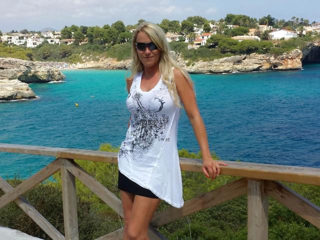 Blonde KIeineDrecksau-hot in white tank top with black sketch, black shorts, sunglasses, standing on wooden railing, blue ocean, rocky coastline, and green hills in background.
